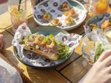 En sandwich med stegt pålæg, grønt og syltede løg ligger på mønstret papir i en skål, omgivet af tallerkener med appetitvækkere og drikkevarer på et træbord i sollys.