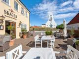 Udendørsservering med hvide borde og stole på Skagen Harbour Hotel, med en hvid kirke og blå himmel i baggrunden.