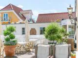 Solrig udendørs terrasse med hvide borde og stole, potteplanter, kurvemøbler og et hvidt hegn foran maleriske huse med røde tegltage.