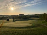 En golfbane ved solopgang med lange skygger, grønt græs, spredte træer og en klar himmel med solen lavt i horisonten.
