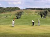 Tre mænd spiller golf på en grøn bane; en holder flagstangen, en gør klar til at putte, og en anden står i nærheden. Træer og bølgende bakker er synlige i baggrunden under en blå himmel.