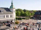 Et livligt bytorv med folk, der går og sidder ved udendørs borde, ved siden af en historisk bygning med et højt spir og omgivet af grønne træer på en solrig dag.