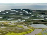 Luftfoto af en snoet flod, der løber gennem et grønt moseområde, med forskellige kanaler og vandområder, der fører til en stor vandmasse i baggrunden.