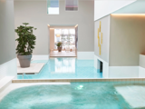 Moderne indendørs poolområde med rene linjer, potteplanter, stærkt naturligt lys og minimalistisk indretning; vandet flyder mellem forbundne poolsektioner.