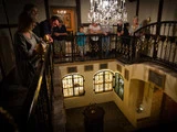 En gruppe mennesker står på en udsmykket balkon med udsigt til et vintage-interiør med lysekroner og store glasmosaikvinduer.