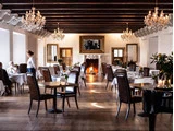 Elegant restaurant med lysekroner, spejle, en tændt pejs og mørke læderstole. En servitrice serverer for gæsterne ved borde med hvide duge og blomster.