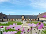 Lyserøde blomster i forgrunden med en gårdhave med et springvand i midten, hvide bænke og en traditionel bygning med spidst tag i baggrunden under en blå himmel.