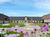En gårdsplads med grønt græs, et centralt springvand af sten, hvide bænke og blomstrende lyserøde blomster i forgrunden, omgivet af traditionelle træbygninger under en blå himmel.