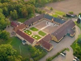 Luftfoto af et stort, gult bygningskompleks i bindingsværk, der danner en firkant omkring en central gårdsplads, omgivet af grønne områder og landbrugsjord.