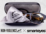Et par skildpaddebriller i et åbent etui med en pudseklud med teksten "NON-VIOLENCE i samarbejde med smarteyes." Non-violence- og smarteyes-logoer er vist nedenfor.