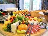 Et stort fad med forskellige oste, pålæg, vindruer, skiveskårne grøntsager og frisk brød er arrangeret på et bord, der er dækket op til buffet i et lyst lokale.