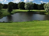 En lille dam omgivet af grønt græs, træer og en golfbane med en sandbunker i baggrunden under en delvis overskyet himmel.