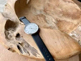 Et minimalistisk armbåndsur med sort læderrem hviler i en stor, udskåret træskål med naturlige teksturer.
