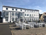 Hvide borde og stole står langs en brostensbelagt gårdhave foran en hvid, treetagers hotel- og restaurantbygning med grå skodder på vinduerne.