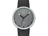 Et minimalistisk armbåndsur med en struktureret grå urskive, grønne timemarkeringer, sort rem og sølvfarvet ramme. Urets visere er sorte med en grøn sekundviser.