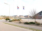 En stille gade med en murstensbygning, to flagstænger med hvide flag, små træer og huse i baggrunden under en lys himmel.