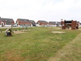 En græsklædt legeplads med gynger, et lille legehus og et stort klatrestativ; huse med røde murstensvægge og hvide kanter ligger i baggrunden.