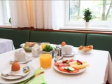 Et morgenbord med juice, æg, brød, ost, pålæg, agurk i skiver og tesæt står ved et vindue med hvide gardiner og en potteplante.