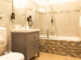 Moderne badeværelse med grå kommode, væghængt hårtørrer, toilet og badekar med glasdøre og stenfliser.