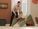 En smilende mand ser på, mens en lille dreng glider ned ad en lille indendørs rutsjebane, mens han holder to bolde i et lyst, hyggeligt rum.