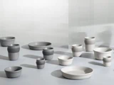 En samling af minimalistiske keramiske skåle og kopper i neutrale grå og hvide toner, arrangeret på en glat overflade med blød belysning og en struktureret lys baggrund.