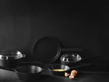 Et sæt sort køkkengrej, herunder en stegepande med æg, gryder med glaslåg og en stegepande, med knuste æggeskaller på en mørk overflade og baggrund.