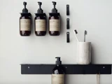 Tre brune sæbedispensere er monteret på en væg over en hylde med en anden dispenser og en hvid kop med to tandbørster.