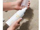 En person holder en hvid flaske Meraki SPF 30 solcreme og tager hætten af, mens der er sand i baggrunden.