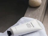 En tube Meraki bodylotion ligger på et hvidt håndklæde ved siden af en rund sten på en betonflade.