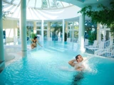 En mand nyder en vandstrålemassage i en indendørs pool, mens en kvinde går ned ad en trappe i vandet under en lys glaskuppel med træer og liggestole i nærheden.