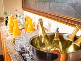 Flasker med mousserende vin i en isspand står ved siden af rækker af tomme glas og kander med orange og gul juice på en bordplade.