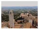 Luftfoto af en gammel italiensk by med stentårne, terrakottatage og bølgende grønne bakker i baggrunden under en grå himmel.