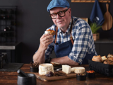 En smilende mand med briller og blå kasket, iført ternet skjorte og forklæde, holder ost og brød ved et rustikt bord med forskellige oste og snacks.