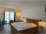 Et lyst hotelværelse med en stor seng, sennepsgult hovedgærde og stole, et skrivebord, lamper og et stort vindue med gennemsigtige gardiner og mørkeblå forhæng.
