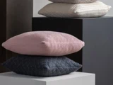 Fire teksturerede puder i pastelfarver og neutrale toner er stablet på moderne firkantede piedestaler mod en ensfarvet baggrund.