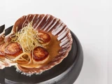 Stegte kammuslinger serveret i en stor muslingeskal, toppet med porrer i tynde skiver og anrettet i en cremet appelsinsauce på et sort fad.