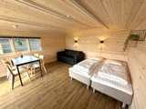 Et hyggeligt værelse med lyse trævægge og -gulve, med to enkeltsenge, en sort sofa, et spisebord med stole, væglamper og en lille plante på en hylde.
