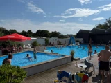 Udendørs pool med folk, der svømmer, leger og slapper af. Børnene bruger en blå rutsjebane, de voksne sidder under en rød parasol, og himlen er delvis overskyet.