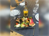 En sort bakke med forskellige appetitvækkere og oste står på et mørkt bord omgivet af tomme vinglas, hvide tallerkener og en flaske mousserende hvidvin.
