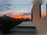 En træterrasse ved siden af et moderne hus ved solnedgang, med levende orange og pink skyer på himlen og grønne planter langs stien.