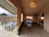 Interiør i et glampingtelt med spisebord, stole, hylder, en seng, der er delvis synlig, og en udendørs terrasse med flere siddepladser og et bord, alt sammen på trægulv.