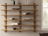 Minimalistiske væghylder i træ med fire vandrette rækker, hvor der står et par skåle, bøger, glas og en lille vase ved siden af et vindue, hvor solen skinner ind.