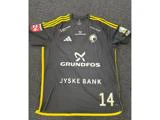 En mørkegrå sportstrøje med gule kanter, nummer 14 og logoer fra Adidas, Grundfos, Jyske Bank og andre sponsorer lå fladt på et gråt gulv med struktur.