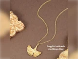 En guldbelagt halskæde med et vedhæng af ginkgoblade vises på en brun baggrund med udsmykket gulddekor. Den danske tekst lyder: Forgyldt halskæde med Gingo blad.
