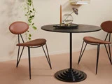 Et moderne rundt sort bord med et guldlys, en glasskål og to brune stole med metalben; en hængende plante i baggrunden.