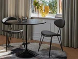 Et rundt sort bord med to sorte stole står på et mønstret tæppe nær et vindue med beige gardiner og en glasvase med blomster i vindueskarmen.
