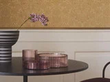 En stribet vase med lilla blomster og to lyserøde glasbeholdere står på et rundt sort bord mod en beige mønstret væg med hvide paneler.