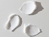 Tre udstrygninger af hvid creme eller lotion på en struktureret lysegrå overflade, set ovenfra.