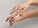 En person smører en klat hvid lotion på håndryggen ved hjælp af fingrene på den anden hånd mod en almindelig baggrund.