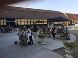 Folk spiser udendørs på en café med store parasoller, borde og stole i en gårdhave ved siden af en bindingsværksbygning på en solrig dag. Serveringspersonalet betjener kunderne.